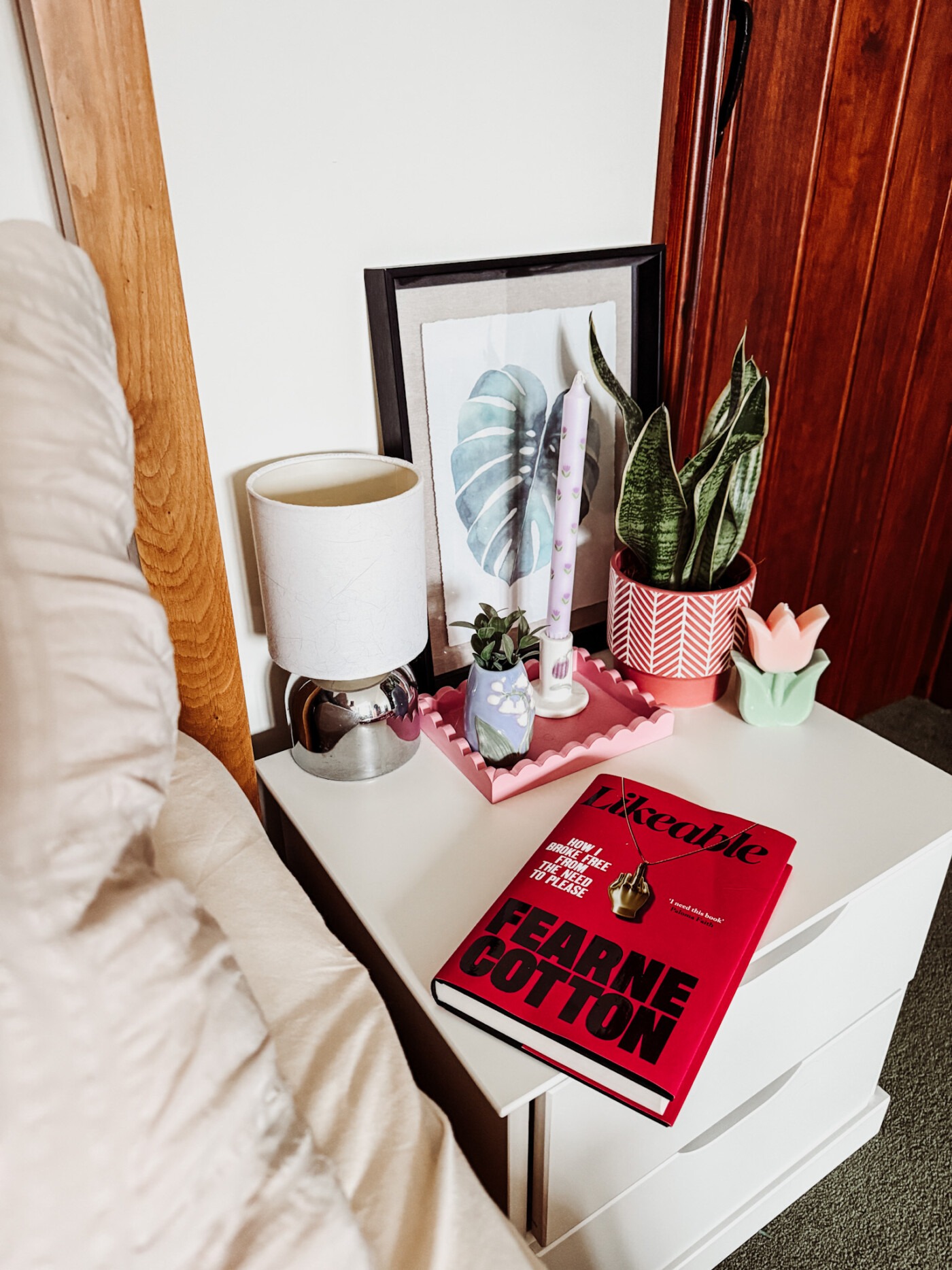 Styled bedside table with potted plant, faux flowers in glass vase, candles on tray, and framed print leaning against white wall