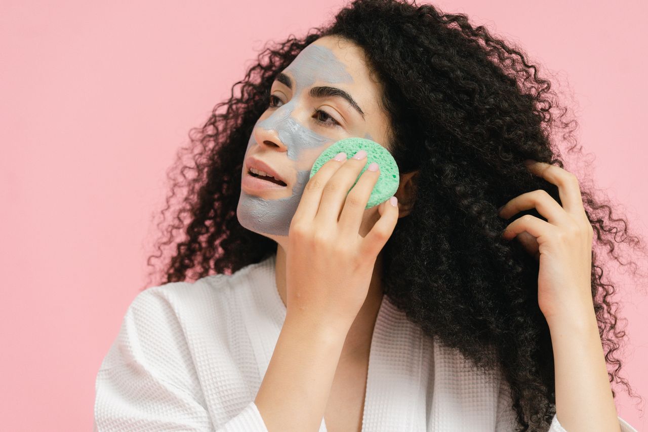 Adult woman with curly hair applying a facial mask using a sponge in a studio setting.